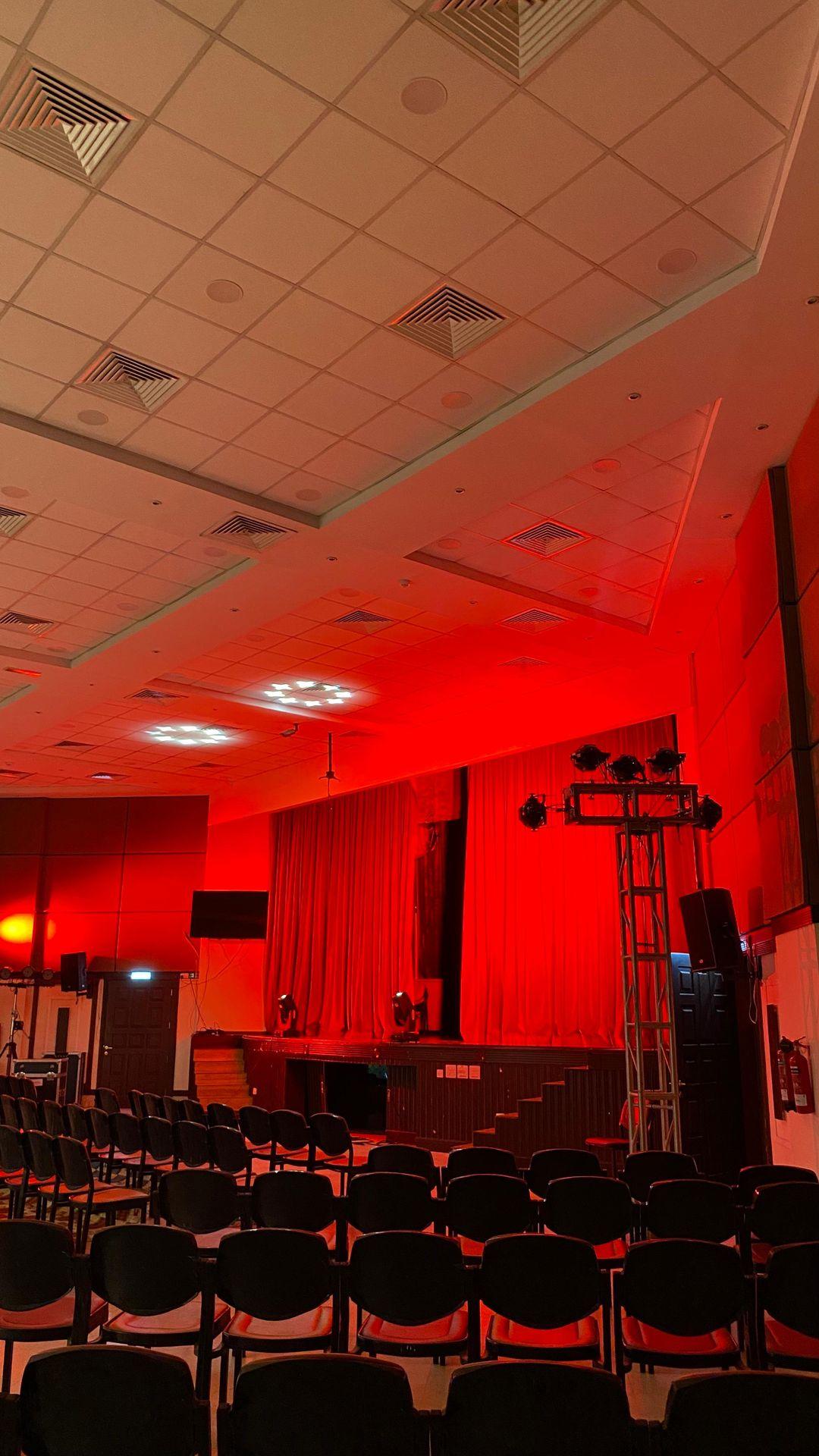 Empty theater with rows of black chairs facing a stage with red curtains and dramatic red lighting.